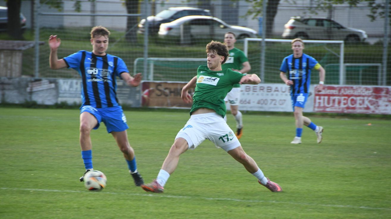 A soccer game is in progress, with players in green, blue, and white uniforms. A player in green kicks the ball while another player in blue tries to block him. A fence with advertisements is behind them.