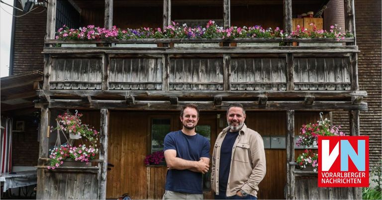 Two men stand in front of a wooden building with a balcony adorned with pink and purple flowers.