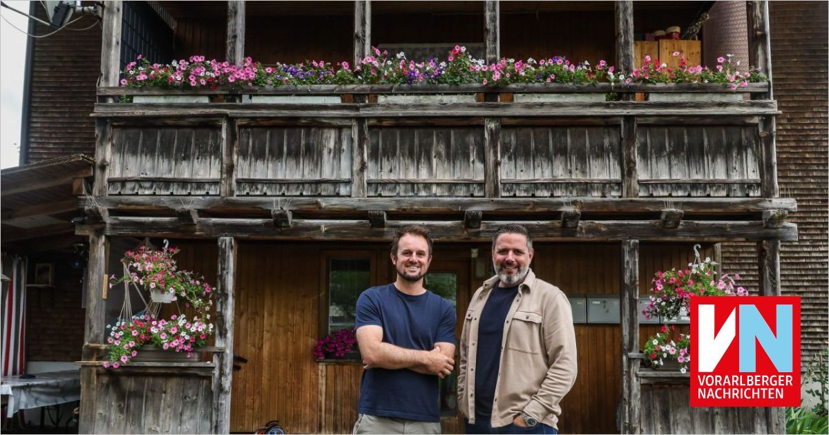 Two men stand in front of a wooden building with a balcony adorned with pink and purple flowers.