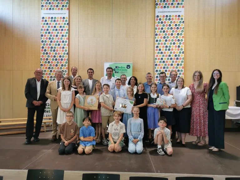 Eine Gruppe von Menschen, einschließlich Erwachsener und Kindern, posiert für ein Foto auf einer Bühne. Einige halten Urkunden und Geschenke. Banner mit dem Text 'Kindergemeinde Rat' sind dahinter.
