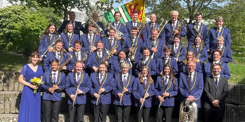 A band with men and women in blue uniforms pose for a picture, holding various musical instruments. Behind them are trees and a flag.