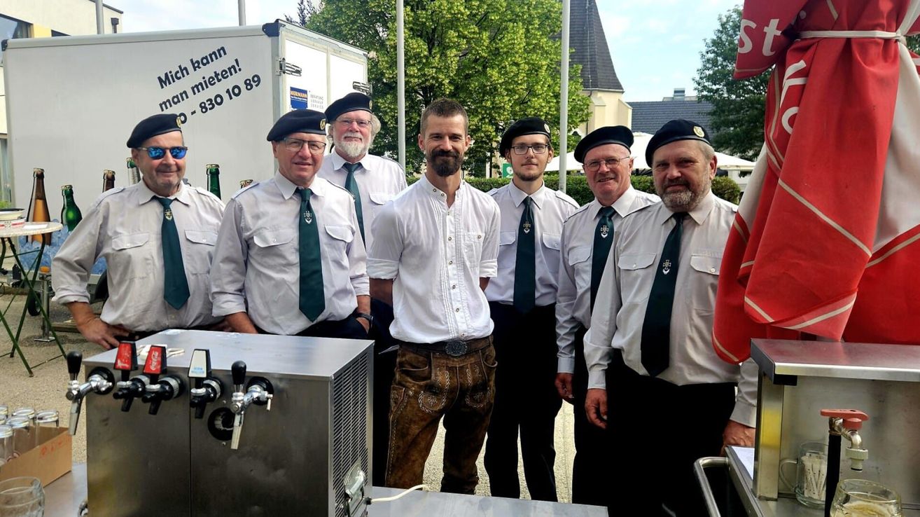 Eine Gruppe von Männern mit grünen Krawatten und Mützen, die neben einem Bierhahn und einem weißen Lastwagen mit einer Nummer darauf stehen.
