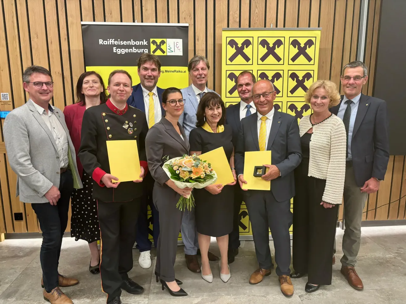 A group of people in formal attire stand in front of a yellow banner with a Raiffeisenbank logo, holding certificates and flowers. A woman in a black dress holds a bouquet of flowers.