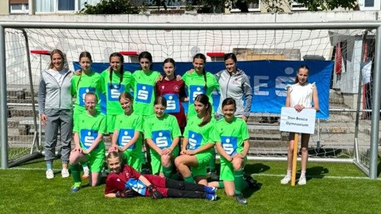 Eine Gruppe junger weiblicher Fußballspielerinnen in grünen Trikots posiert für ein Foto vor einem Fußballtor. Eine Frau in einer grauen Jacke steht hinter ihnen.