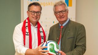 Zwei Männer stehen dicht beieinander und halten einen Fußball. Der Mann auf der linken Seite trägt eine Schürze und ein weißes Hemd. Der Mann auf der rechten Seite trägt eine grüne Jacke und eine rote Krawatte.