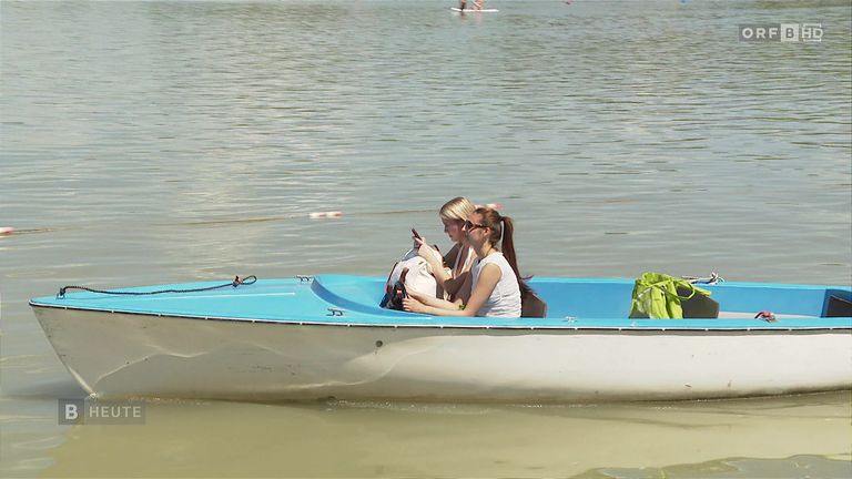 Zwei Frauen sitzen in einem blau-weißen Boot auf ruhigem Wasser, eine hält ein Handy und die andere schaut etwas an. Eine grüne Tasche liegt am Bootsrand.