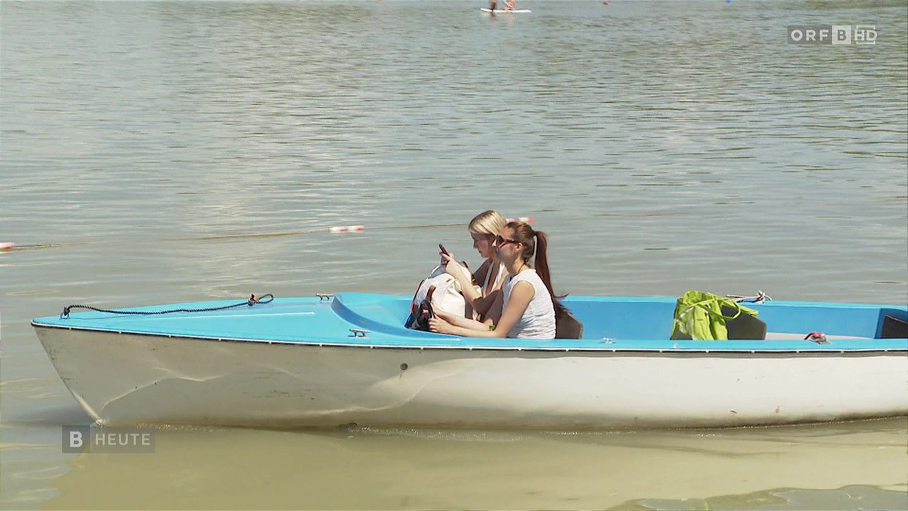 Zwei Frauen sitzen in einem blau-weißen Boot auf ruhigem Wasser, eine hält ein Handy und die andere schaut etwas an. Eine grüne Tasche liegt am Bootsrand.