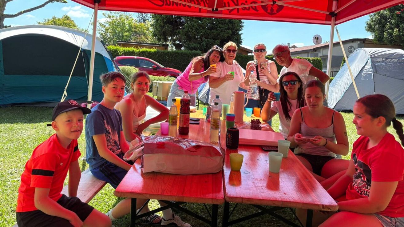 A group of people, including children and adults, are gathered around a picnic table with drinks and food. They are under a red tent in a park.