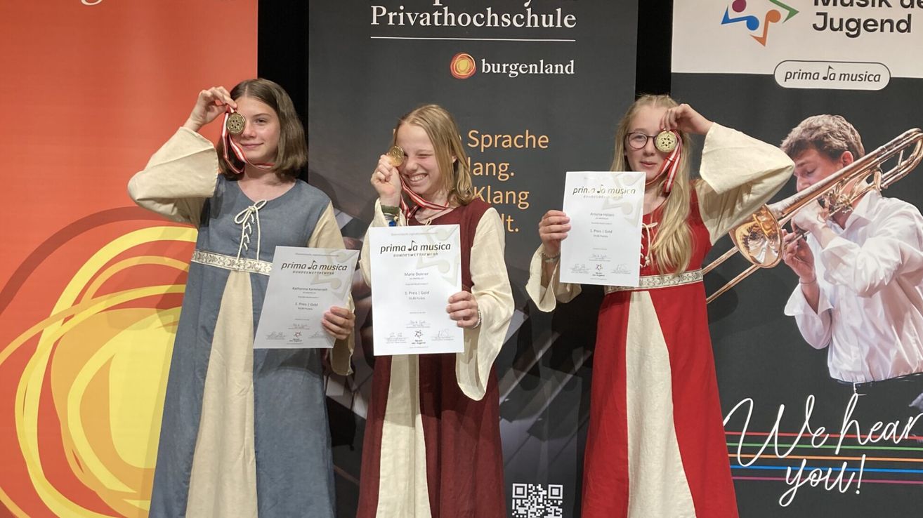 Three young women in medieval-style costumes are holding awards and certificates. They are standing in front of a backdrop with text and logos.