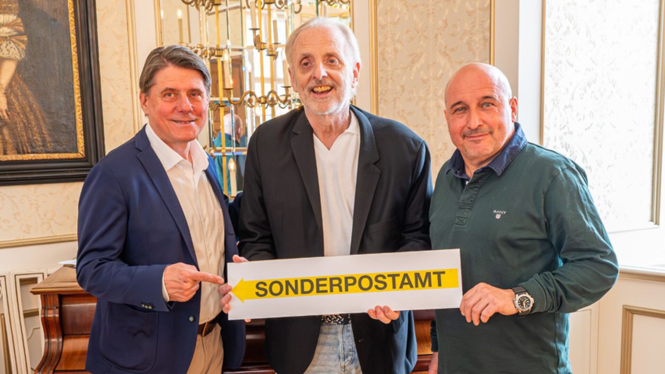 Three men stand next to each other, smiling. One man holds a sign that reads 'SONDERPOSTAMT'. They appear to be posing for a photograph in a formal setting.
