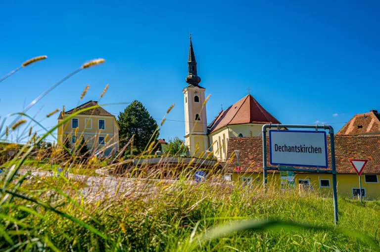 Die Dechantskirche erhebt sich mit einem Turm, einer Uhr und einem roten Dach in einem Dorf. Ein Schild steht Dechantskirche. Eine Brücke und Bäume sind im Vordergrund.