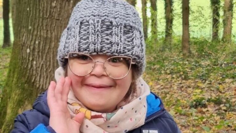 A young girl wearing glasses and a gray knit beanie waves in a forest. She wears a polka-dot scarf and a blue jacket with a white logo.