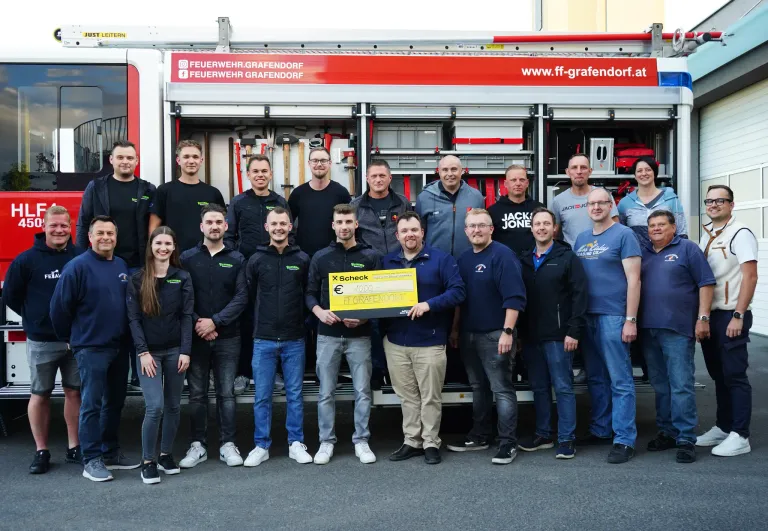 Eine Gruppe von Feuerwehrleuten steht vor einem Feuerwehrauto, einer hält einen Scheck. Sie sind alle in Uniform gekleidet. Das Feuerwehrauto zeigt die Website-Adresse an.