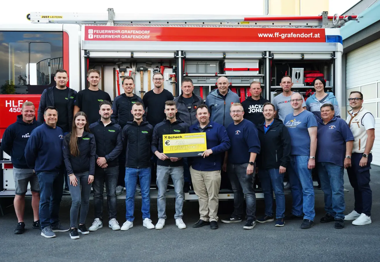 A group of firefighters stands in front of a fire truck, with one holding a check. They are all dressed in uniform. The fire truck displays the website address.