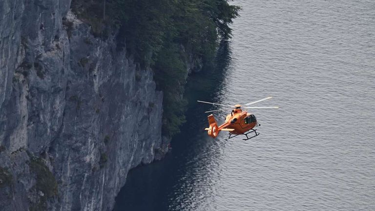 An orange helicopter is flying near a steep cliff and water body with a clear sky.