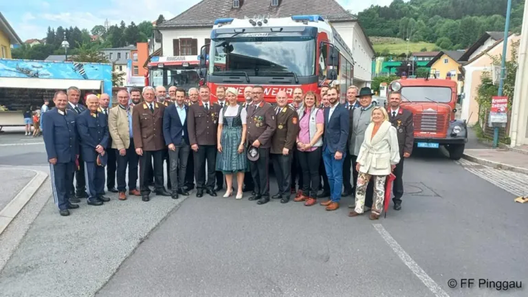 Eine Gruppe von Menschen in Uniformen steht vor einem Feuerwehrauto im Freien. Das Feuerwehrauto hat Text darauf.