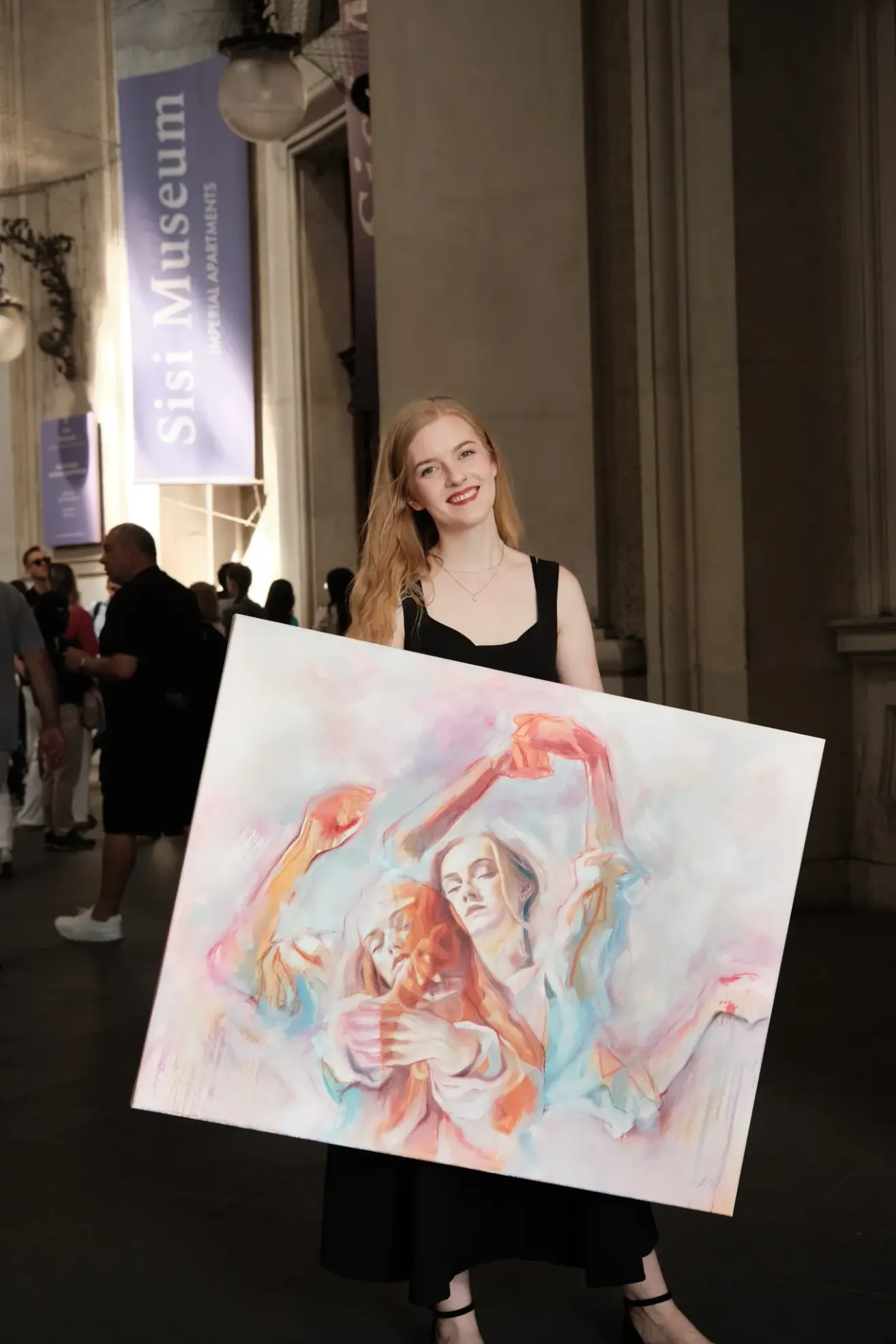 A woman holds a large painting of two women embracing at an event, with people in a hallway in the background.
