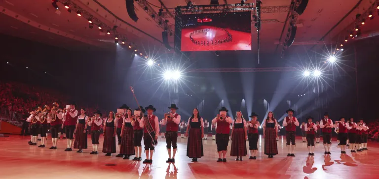Eine Gruppe von Musikern in traditioneller Kleidung tritt auf der Bühne auf, mit einer großen Leinwand im Hintergrund.
