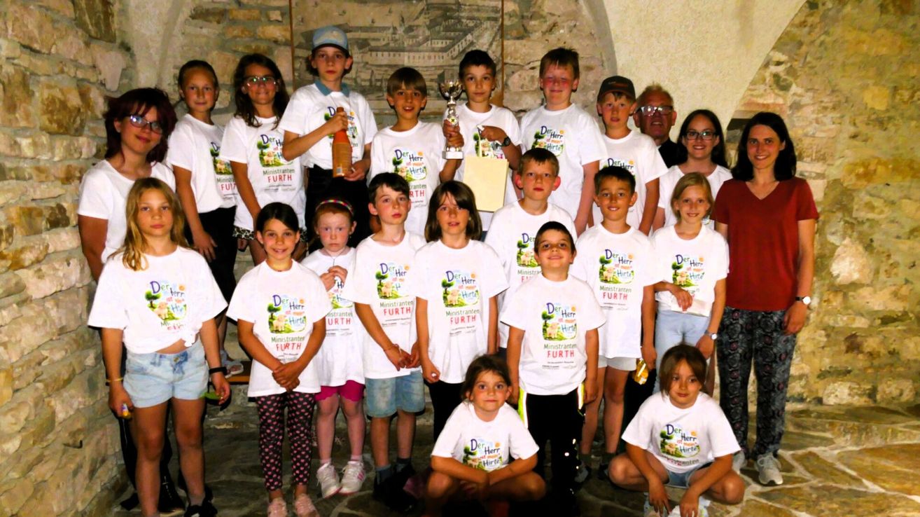 Eine Gruppe von Kindern, die weiße T-Shirts mit dem Logo 'Der Herr' und verschiedenen Accessoires tragen, posiert für ein Foto, einige halten Pokale.