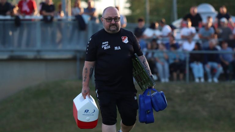 A man in a black sports jersey with a white logo and text walks on a field, carrying two bags. In the background, there are people watching.