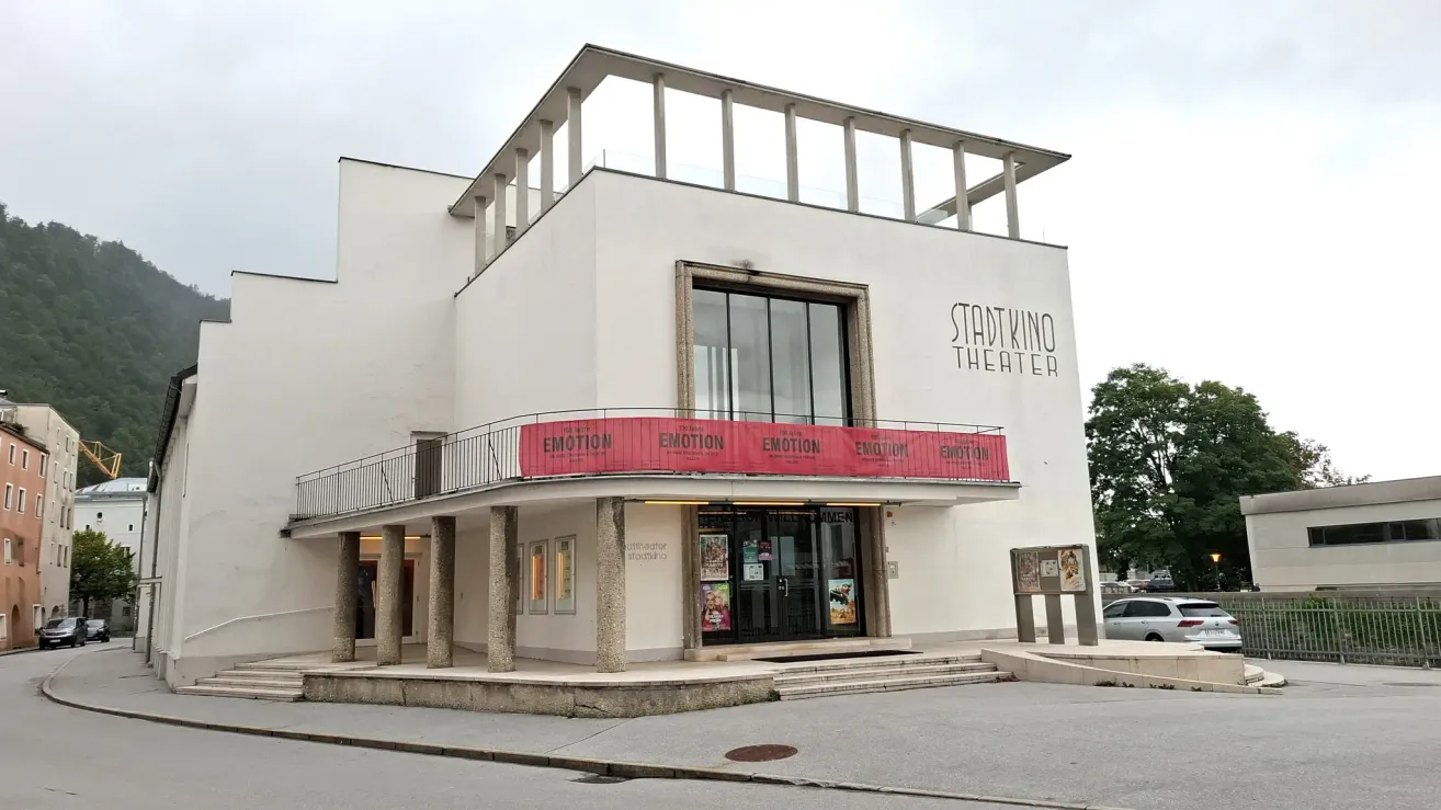 Außenansicht eines modernen, weißen Gebäudes mit einem roten Banner, das 'Emotion' über dem Eingang zeigt. Es hat ein Glasfenster und Türen, umgeben von Säulen. Ein Balkon befindet sich über dem Eingang mit Plakaten darunter.