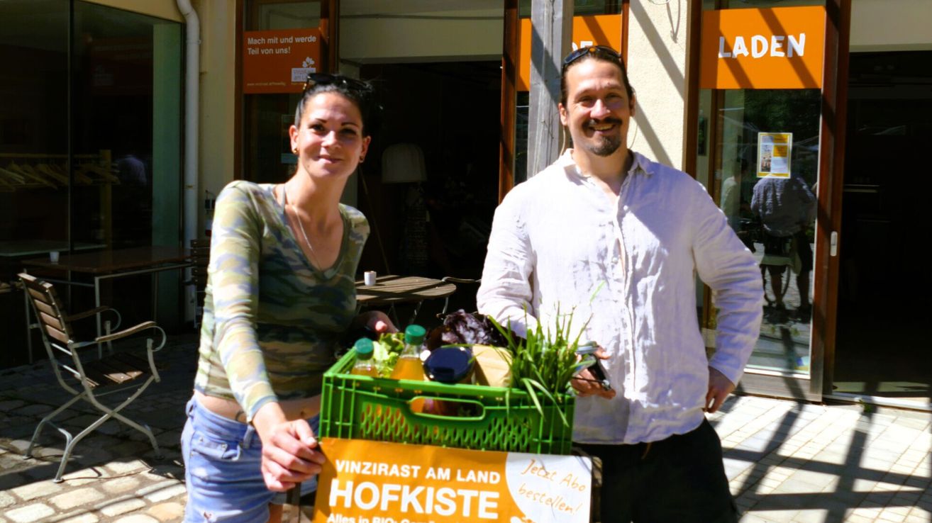 Ein Mann und eine Frau stehen neben einem grünen Korb voller Produkte. Die Frau hält ein Schild mit der Aufschrift 'VINZIRASAM LAND HOFKISTE'. Sie lächeln beide und schauen in die Kamera. Dahinter befindet sich ein Geschäft mit orangefarbener Beschilderung.
