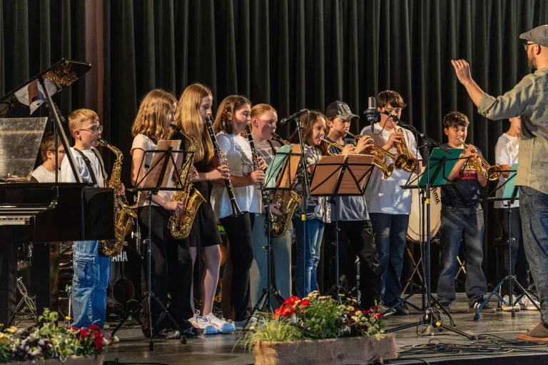 A group of young musicians perform on stage with various instruments like saxophones and trumpets. They are standing in front of a black backdrop and a flower arrangement.