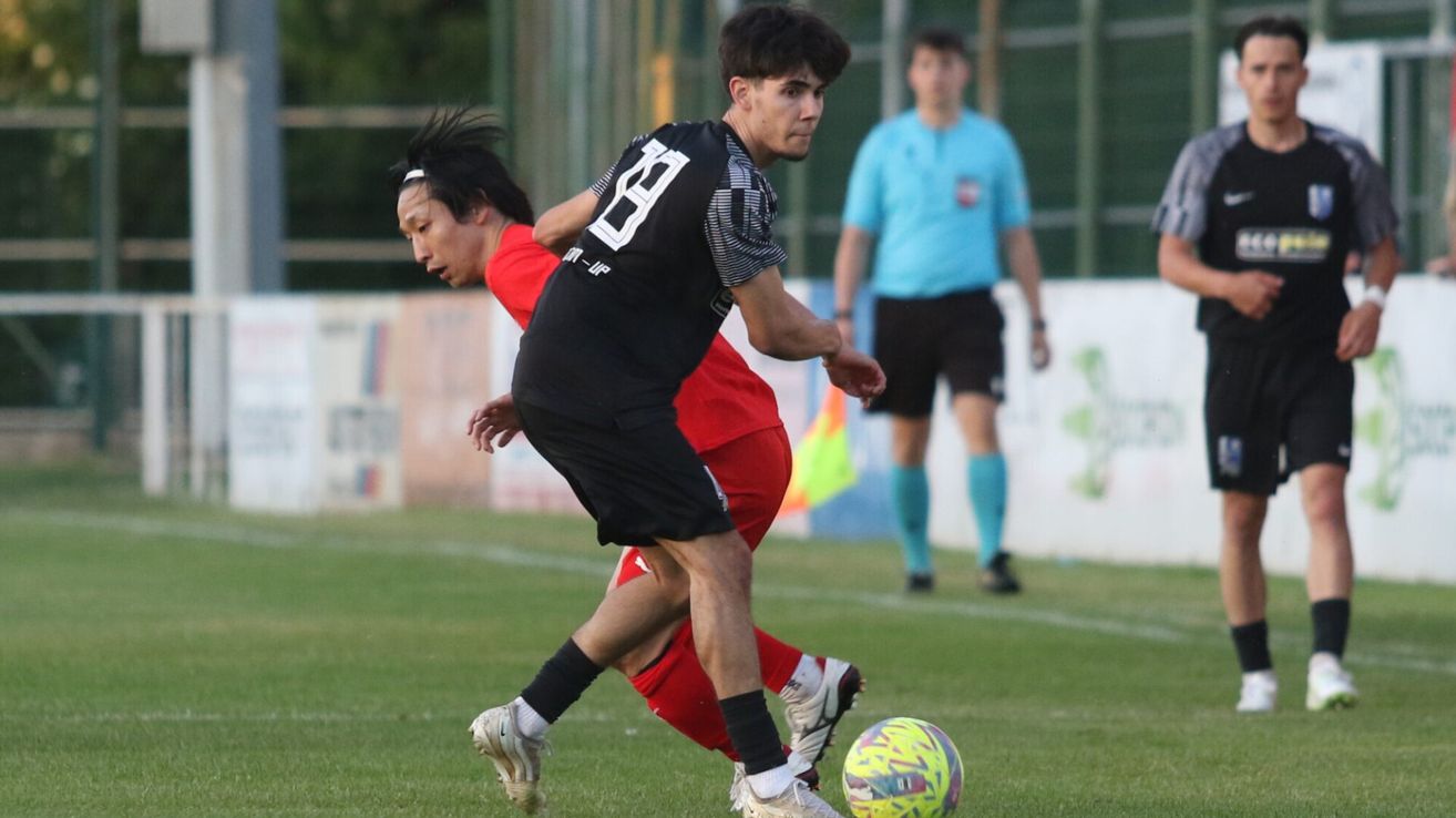 A soccer player in a black jersey chases another in a red jersey. A referee stands behind them. A soccer ball lies on the ground.