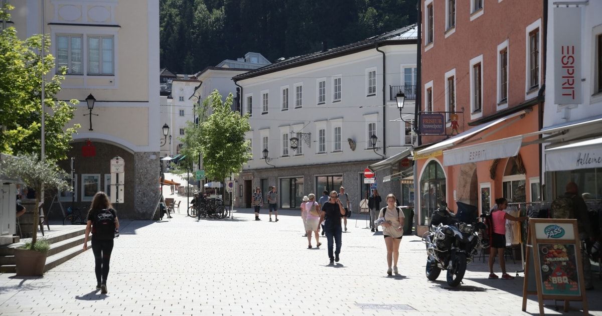 Menschen schlendern eine gepflasterte Straße entlang, gesäumt von Gebäuden, geparkten Fahrrädern und Bäumen und Straßenlaternen. Eines Motorrad ist geparkt an der Seite.