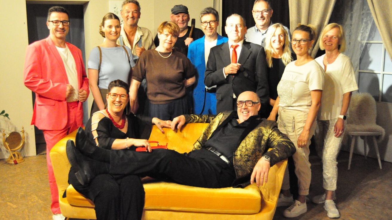 A group of people in a room, some sitting on a couch. A man in a black and gold jacket is sitting on the couch.