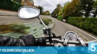 A motorcycle view from the handlebars showing a curved road with bushes and houses on a cloudy day.