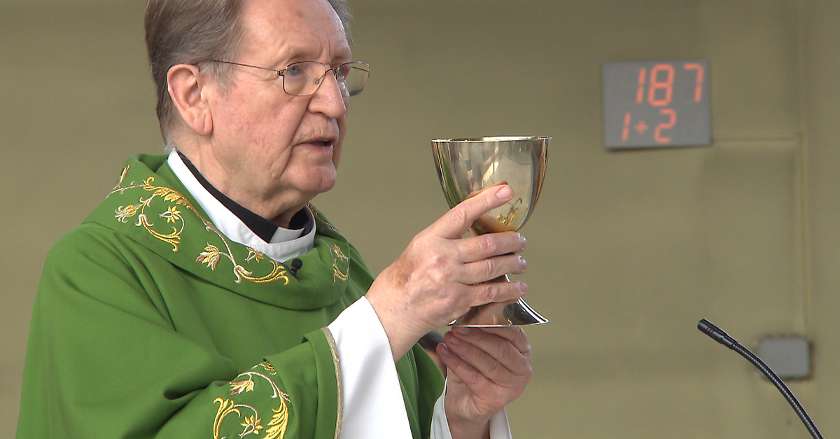 Ein älterer Priester in grüner Robe und mit Brille hält einen Kelch. Er scheint zu beten oder zu sprechen. Eine digitale Uhr zeigt 18:02 an.