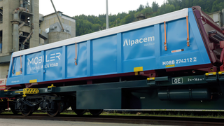Ein blauer Zugwagen mit der Aufschrift Alpacem Austria steht auf einem Gleis, mit einem Wald im Hintergrund.
