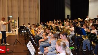 Eine große Gruppe von Kindern in einem Saal, sitzend und verschiedene Musikinstrumente spielend, darunter Trompeten und Flöten. Die Kinder tragen weiße Hemden und Turnschuhe.