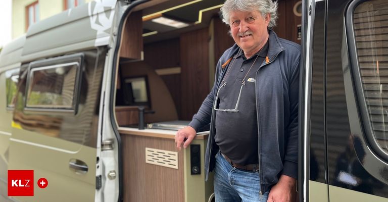 Ein älterer Mann mit einem Lächeln steht in einem Wohnmobil, trägt eine Jacke und seine Brille hängt an seinem Hemd.
