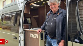 An older man with a smile stands inside a camper van, wearing a jacket and glasses on his shirt.