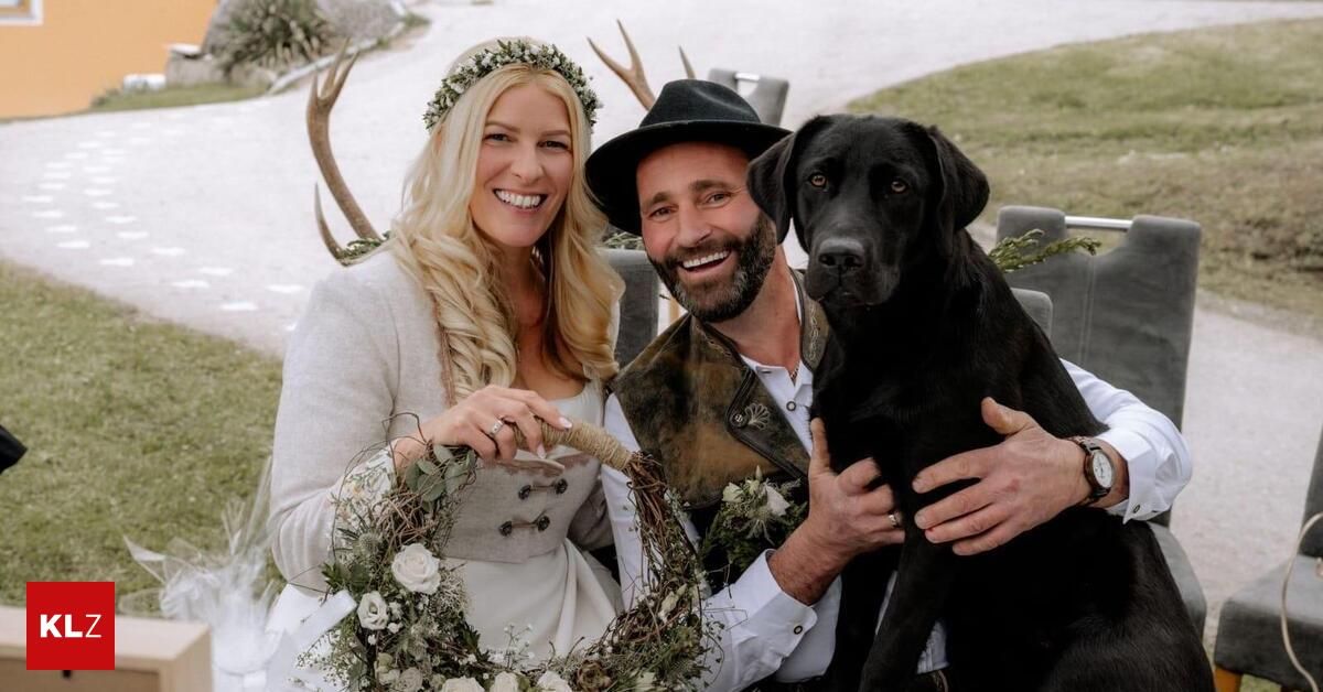 Ein lächelndes Paar an einem Wintertag, das eine Girlande und einen schwarzen Hund hält, mit Hirschgeweihen und grünem Gras im Hintergrund.