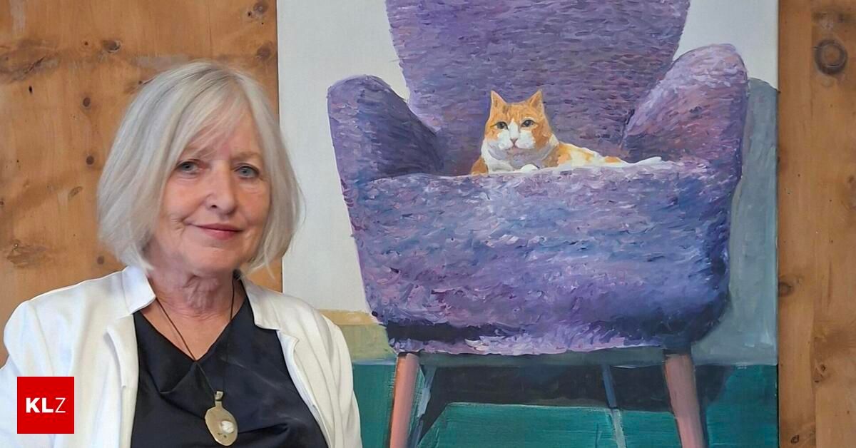 An older woman stands next to a purple armchair with a cat sitting inside. The cat looks relaxed and is wearing a white collar.