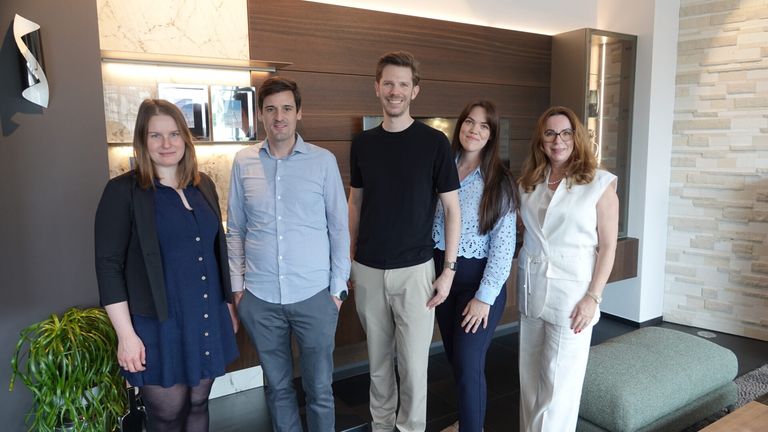 Five people stand together in an indoor area, smiling for a photo. Three men and two women. The man on the left wears a blue shirt, the man in the middle a black t-shirt, and the woman on the right wears a white blouse.