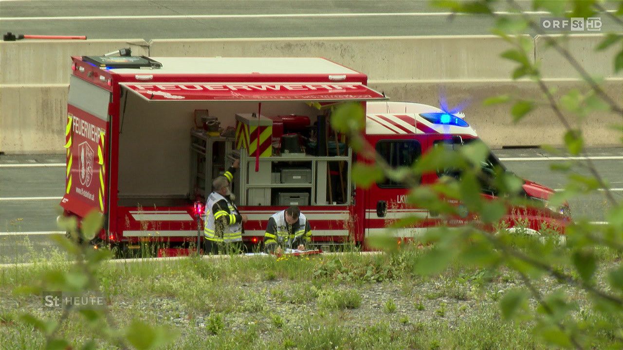 Zwei Feuerwehrleute arbeiten in der Nähe eines Feuerwehrwagens. Einer steht, während der andere sitzt. Der Wagen ist auf einer Straße geparkt.