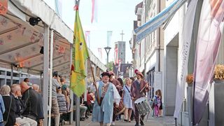 People dressed in historical costumes parade through a narrow street, holding flags and drums, while spectators watch from benches and buildings.