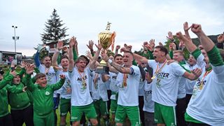 Soccer players celebrate with a golden trophy, wearing white jerseys with green shorts, and several have medals around their necks. They raise their hands in joy under a cloudy sky.