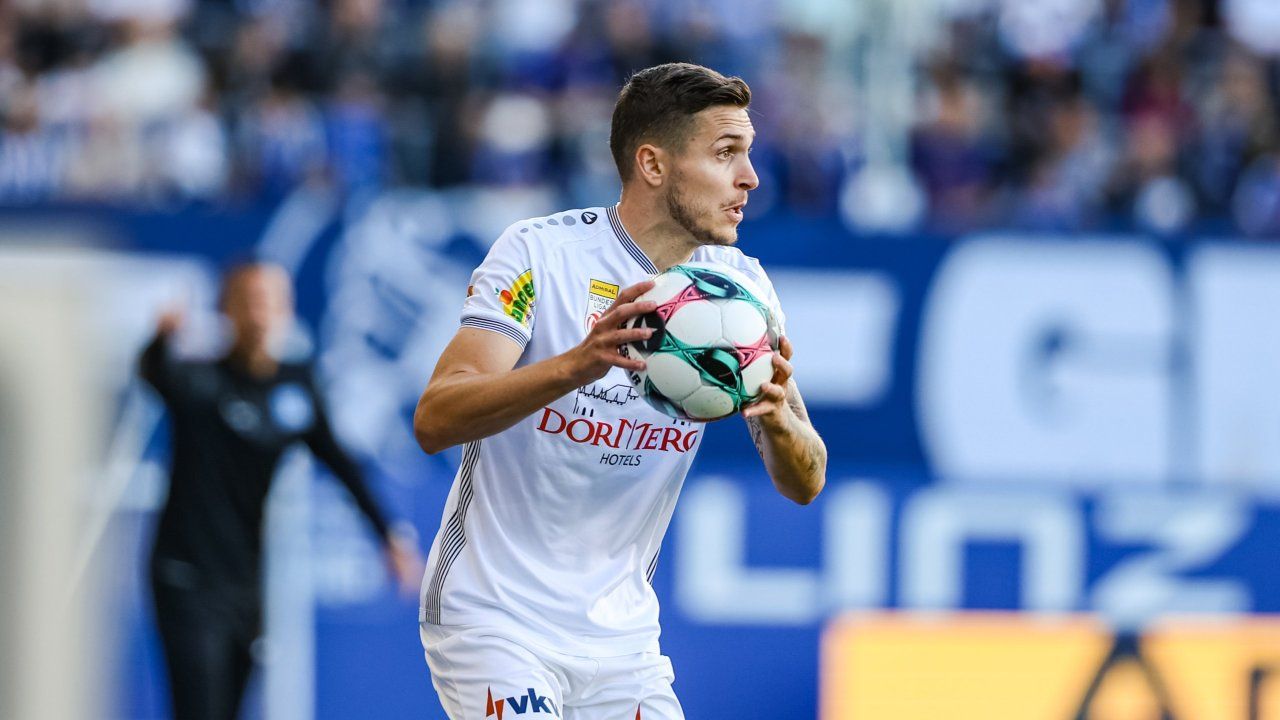 A soccer player runs on the field, holding a soccer ball. He is wearing a white uniform with the word Dorintero Hotels on it.
