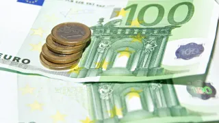 A stack of Euro coins and a 100 Euro bill on a white background. The coins are golden and feature a unique design. The bill has yellow stars and a building in the background.