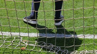 A person in black pants with white stripes is standing on a soccer field with a white net in front of them.