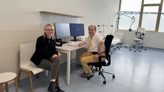 Two people are sitting at a desk in an office room, smiling. The room has a white desk, chairs, monitors, and medical equipment.