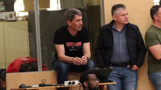 Three men sit on a bench, one in a black shirt with red letters, another in a black jacket. They look serious.