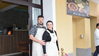 A man and a woman in aprons stand smiling in front of a restaurant.