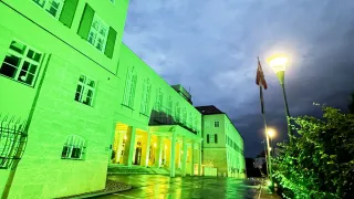 A building with green lights is visible at night. The road is wet and there are streetlights on the side.
