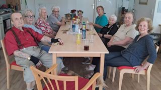 A group of elderly women and men sit around a table in a room. There are glasses, bottles, and snacks on the table. One woman smiles, and others look content. A dog lies on the floor.